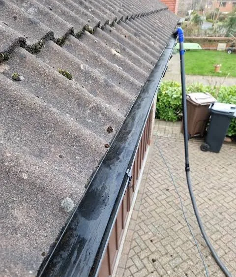 Freshly cleared black uPVC gutter along a tiled roof, with the gutter vacuum hose running down to the driveway